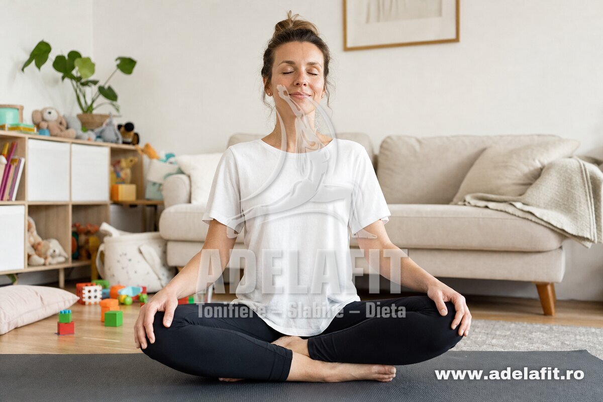 Femeie europeană face yoga acasă, stând în poziție de meditație, zâmbind relaxată.