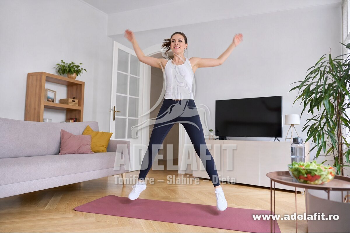 Femeie făcând jumping jacks acasă, pe saltea, cu apă și salată proaspătă pe masă