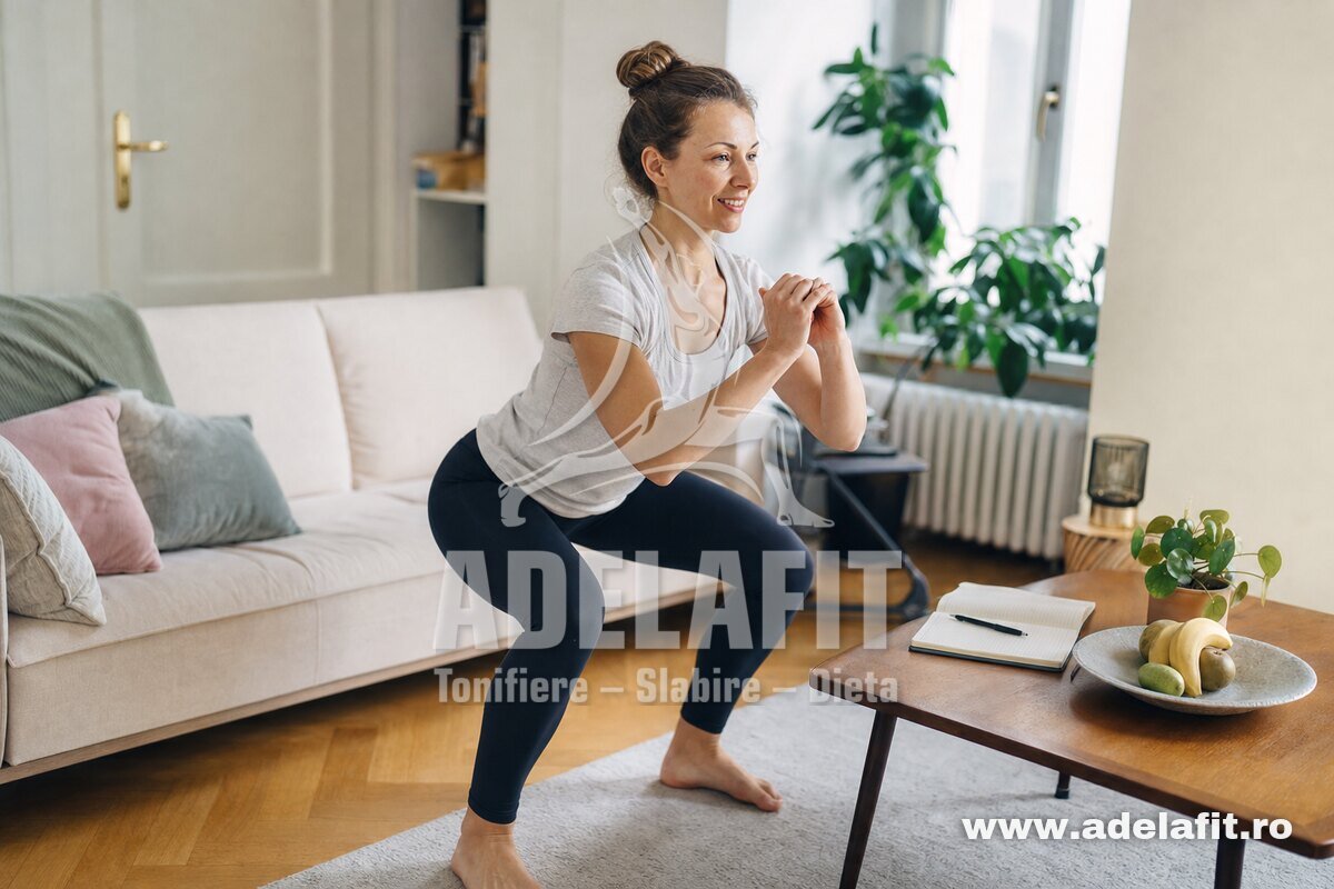 Femeie făcând genuflexiuni acasă, în ținută sport, cu un carnețel pe masă lângă ea