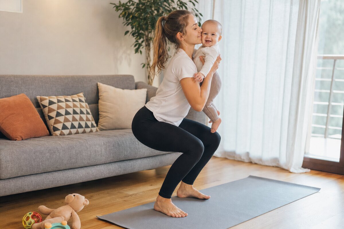 Mamă făcând genuflexiuni cu bebelușul în brațe într-un living, pe covor de yoga